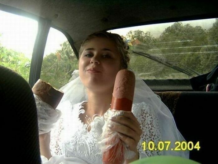 Bride in wedding dress holding two large sausages inside a car, an awkward wedding photo that is both funny and cringe-worthy.