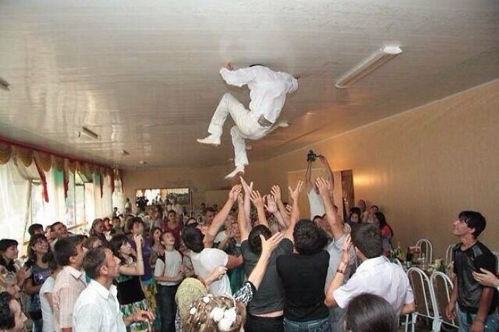 Groom awkwardly caught midair near low ceiling during a lively wedding celebration with guests reaching up.