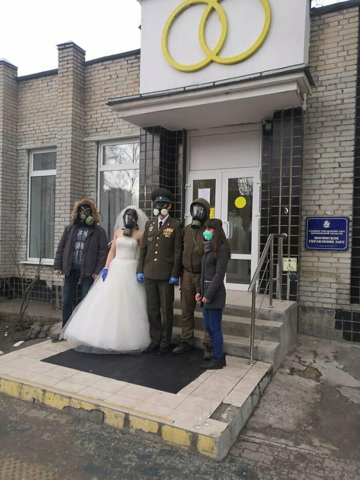 Bride, groom, and guests wearing gas masks and masks in an awkward wedding photo outside a building with ring symbols.