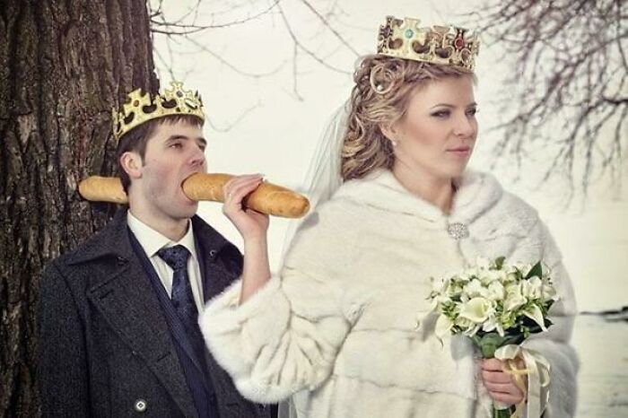 Couple wearing crowns with bride holding baguette near groom’s mouth in an awkward wedding photo outdoors by tree.