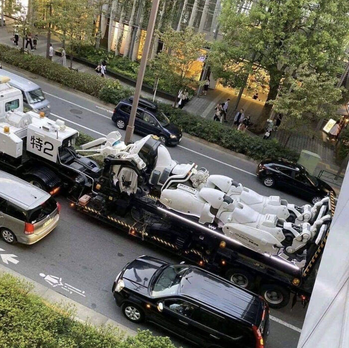 Large truck carrying multiple white robot figures on city street, a confusing picture that is funny and intriguing.
