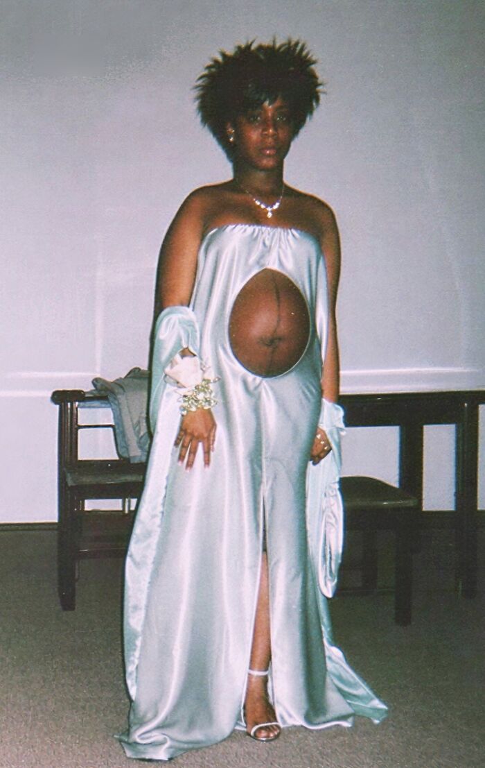 Woman wearing a questionable taste wedding dress with a large cutout revealing her belly, standing indoors near a bench.