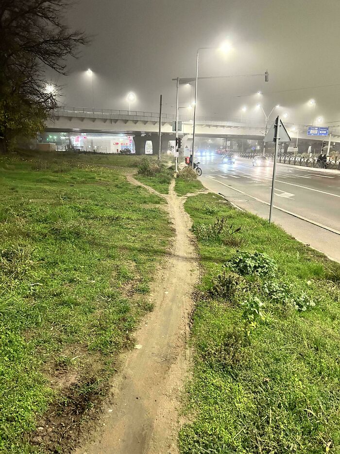 Desire path cutting across grass near a city road and underpass, showing how people walk in reality versus planned routes.