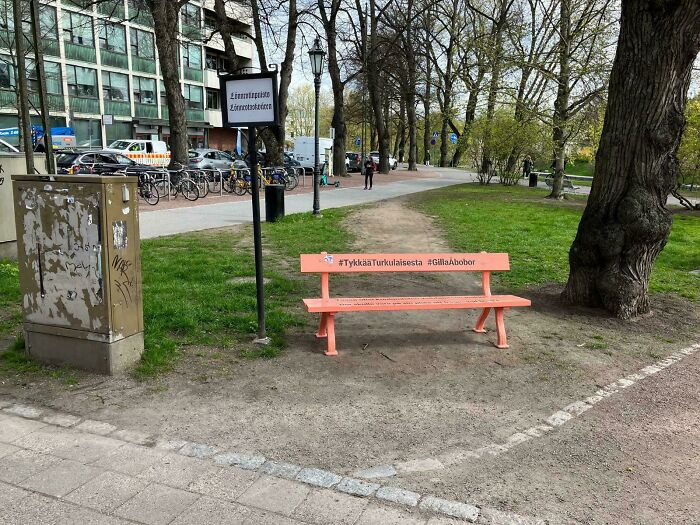 Desire path worn in grass alongside paved walkway near an orange bench in an urban city park area