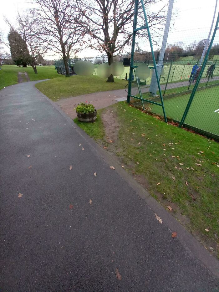 Desire path worn on grass beside paved city walkway near sports field and trees in an urban park setting.