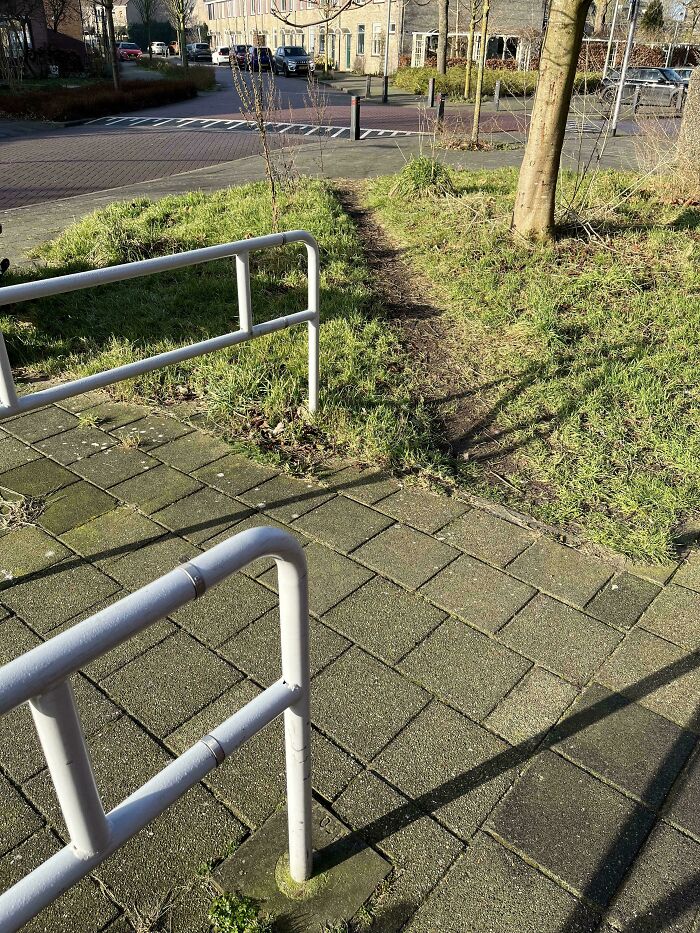 A visible desire path worn through grass beside a paved urban sidewalk and metal railings in a residential area.