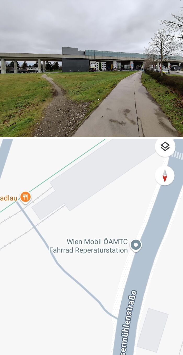 Desire path cutting across grass beside paved walkway near urban area and railway infrastructure in city.