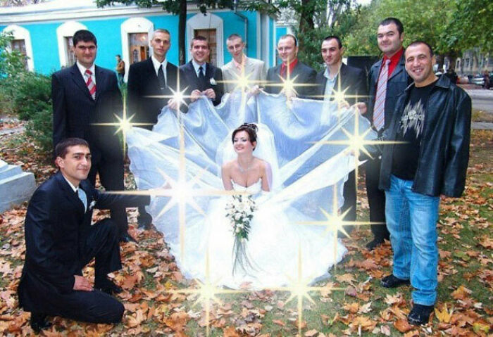 Bride sitting in a wedding dress held by groomsmen outdoors, surrounded by sparkling light effects, awkward wedding photo moment.