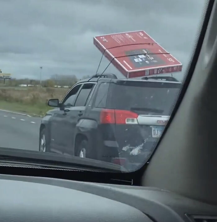 Confusing picture of a large TV box tied awkwardly to the roof of a black SUV on a cloudy day.