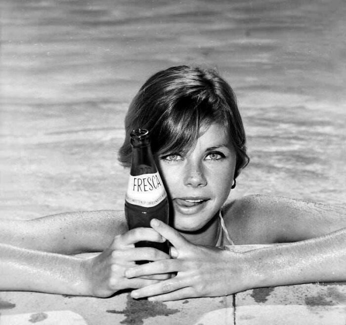 Young woman in vintage ad holding a Fresca bottle while relaxing at the pool, reflecting retro society rules.