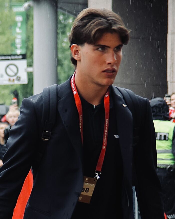 Cesare Casadei wearing a black blazer and red lanyard, walking outdoors with a focused expression in a rainy setting.