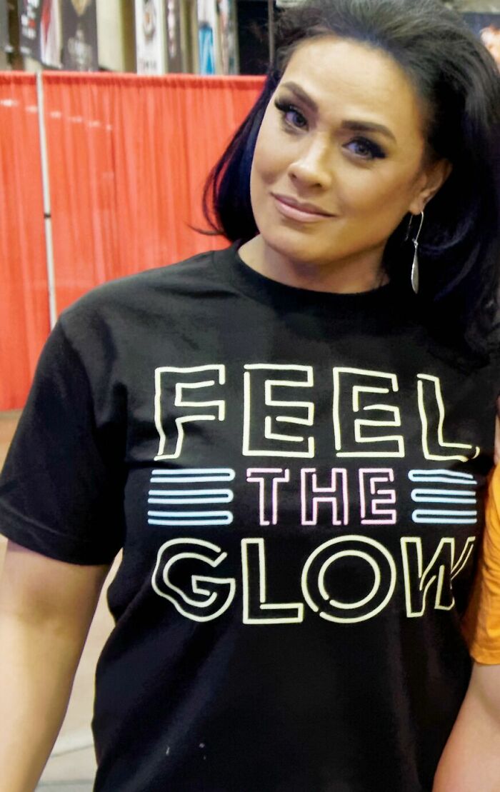 Tamina Snuka wearing a black Feel the Glow shirt with long dark hair and hoop earrings at an indoor event.