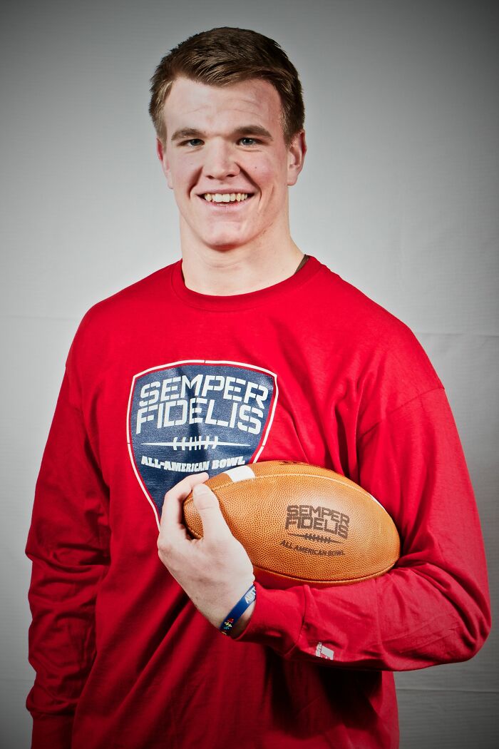 Mike McGlinchey smiling, holding a football, wearing a red Semper Fidelis All-American Bowl shirt in a studio setting.