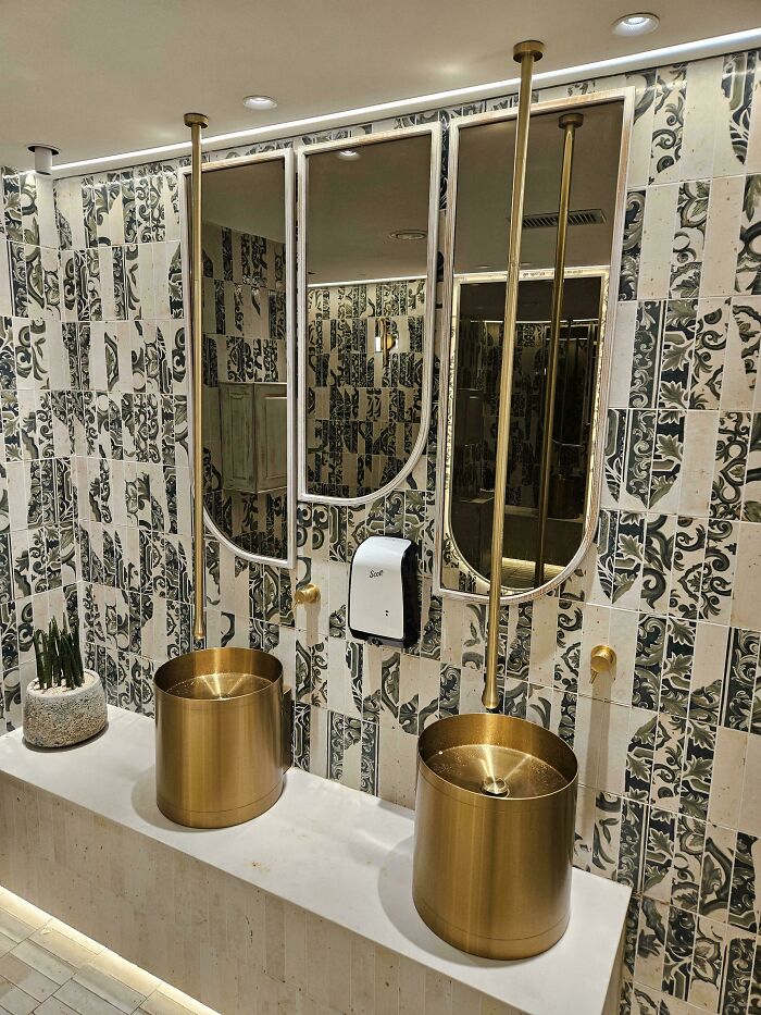 Modern bathroom with gold cylindrical sinks and tall faucets, featuring a patterned tile wall and unusual designer decisions.