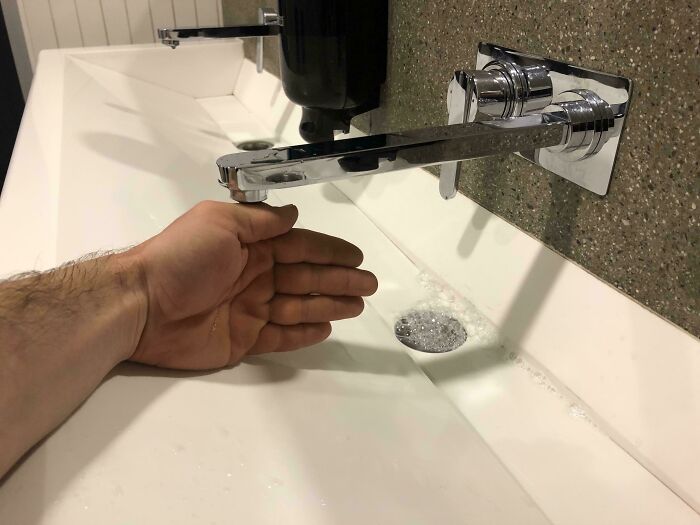 Hand under wall-mounted faucet over a slanted sink with poor drainage, showing a designers dumb decision causing water pooling.