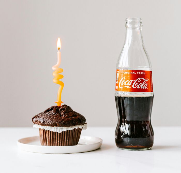 Chocolate cupcake with a lit spiral candle on a plate next to a classic glass bottle of Coca Cola drink on a table