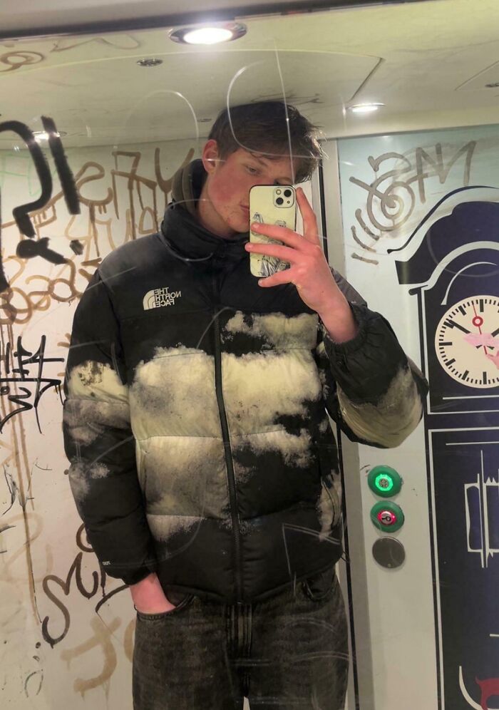 Young tall person taking a mirror selfie in a graffiti-covered elevator, wearing a North Face jacket and holding a phone.