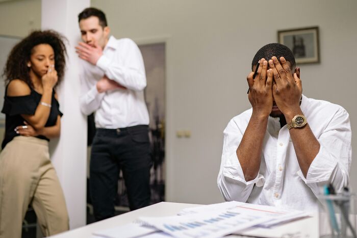 Stressed man covering face at desk while two coworkers whisper nearby, depicting the worst coworkers at work conflict.