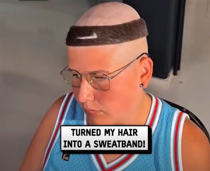 Man with disastrous hairstyle shaved bald except for a thick hair strip designed like a sweatband, wearing glasses and a blue jersey.