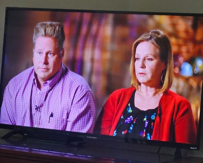 Two people sitting in an interview setting, featuring a man with a disastrous hairstyle and a woman in a red cardigan.