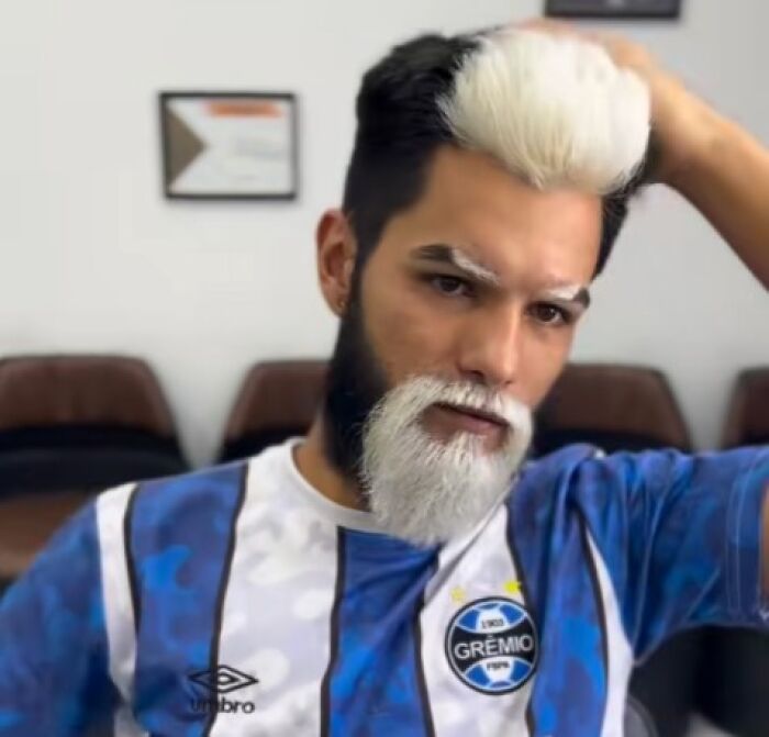 Man with a black and white split dyed hairstyle and beard wearing a blue and white Gremio soccer jersey indoors.