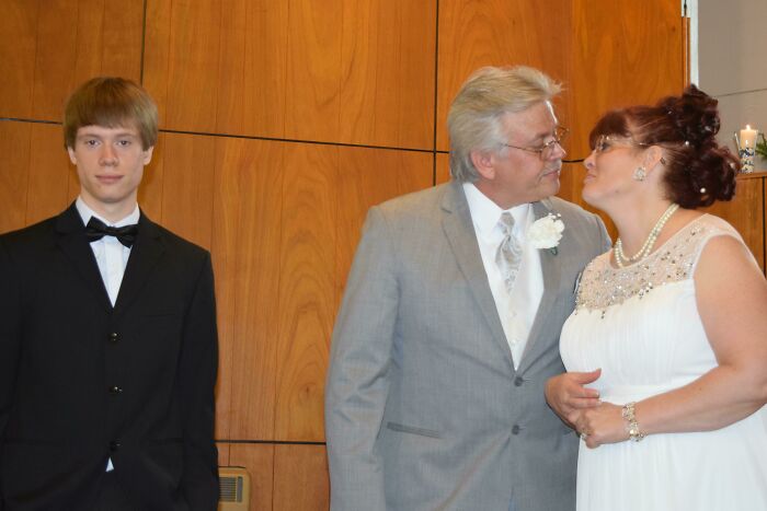 Awkward wedding photo showing a bride and groom leaning in to kiss while a teenage boy in a tuxedo looks uncomfortable nearby.