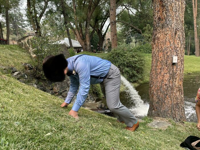 Man in blue shirt awkwardly falling on grass near pond with trees in background, capturing awkward wedding photo moment