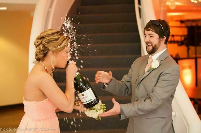 Bride in a pink dress accidentally spraying champagne on groom in a suit, one of the awkward wedding photos caught in action.