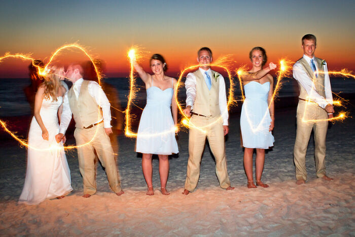 Couples and wedding party on beach at sunset creating love sign with sparklers in awkward wedding photos.