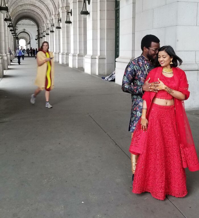 Couple in traditional attire poses awkwardly for wedding photo under arched walkway with passerby in the background.