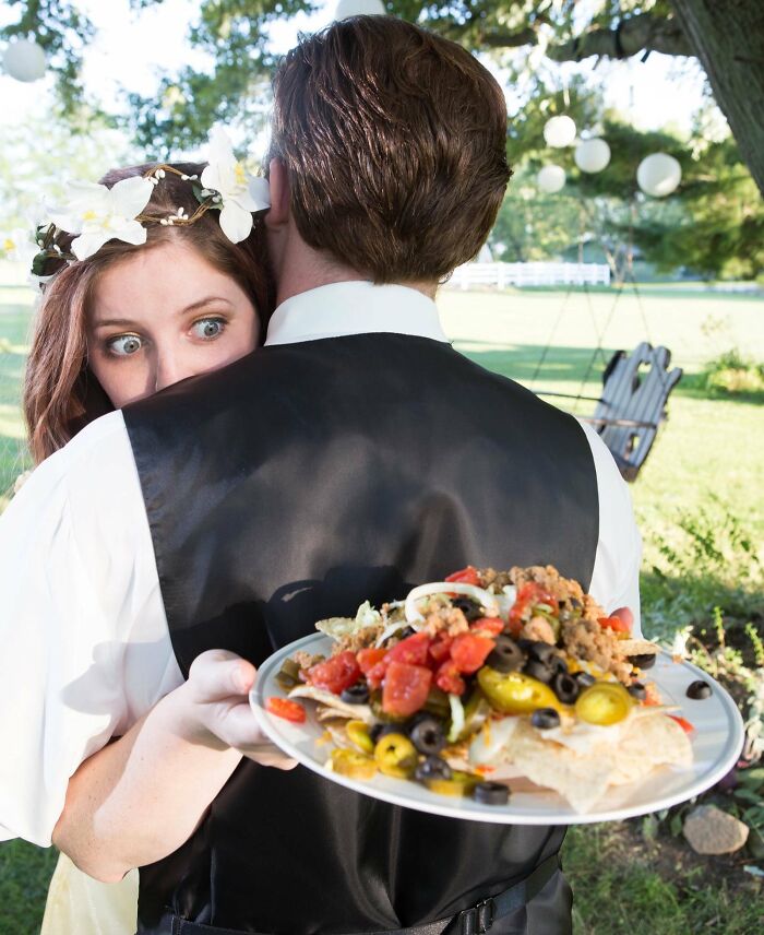 Bride with a shocked expression hiding behind groom holding a plate of nachos in awkward wedding photos outdoors.