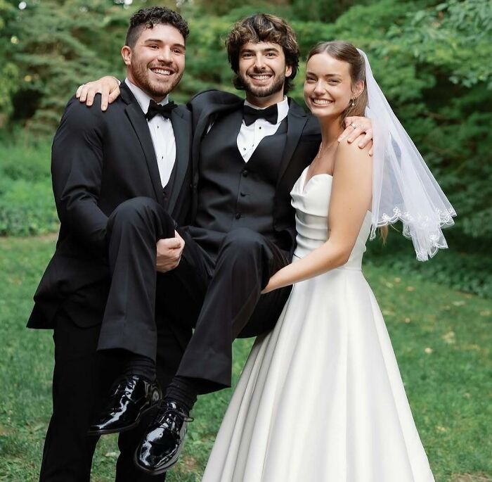 Bride in white gown and veil and two groomsmen lifting another in a tuxedo in awkward wedding photos outdoors.