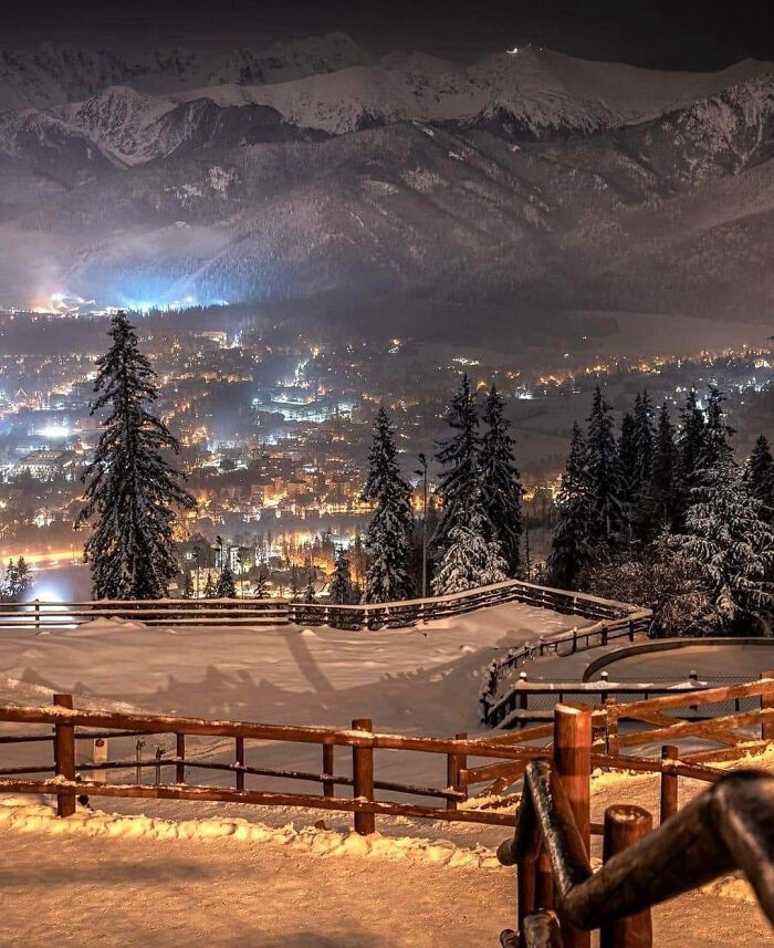 Snow-covered trees and wooden fences overlook a warmly lit town below, capturing cozy winter views this year.
