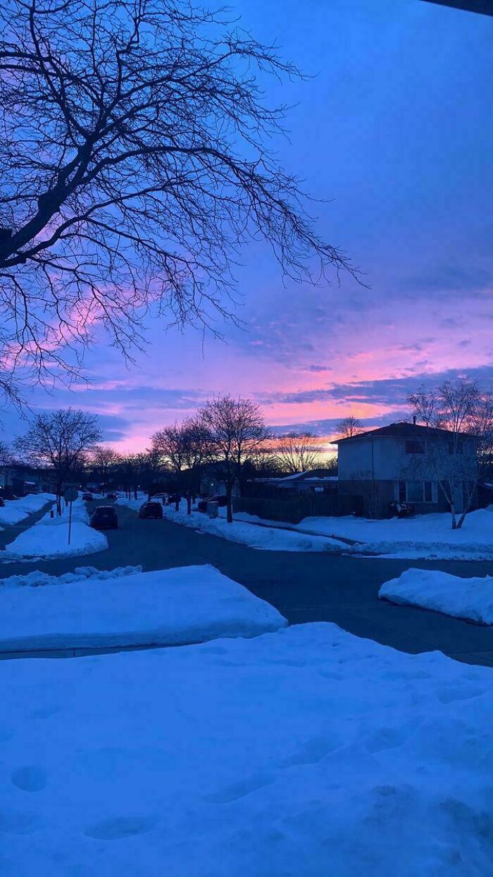 Quiet winter neighborhood street at sunset with snow-covered lawns and bare trees under a colorful sky.