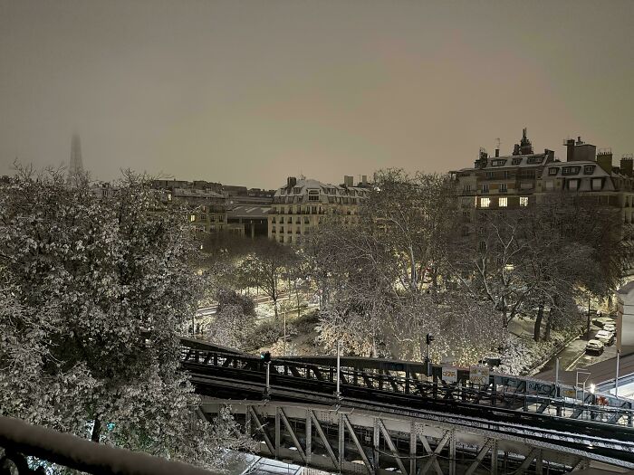 Snow-covered urban park and railway bridge at night creating a cozy winter atmosphere with warm building lights.