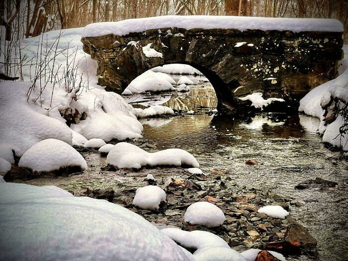 Snow-covered stone bridge over a gently flowing creek surrounded by winter forest, showcasing cozy winter scenes outdoors.