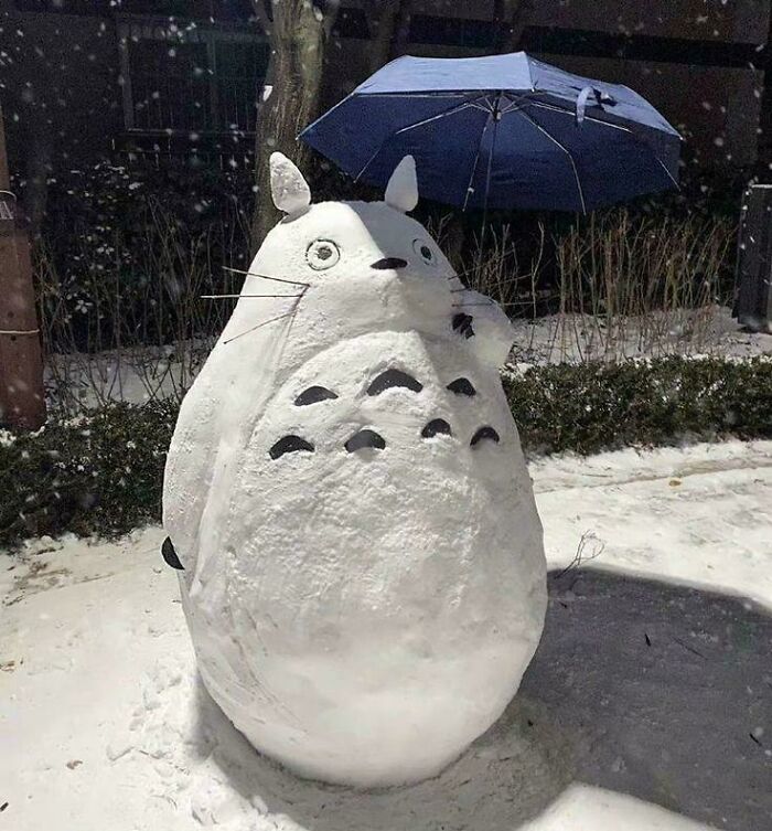 Snow sculpture of Totoro holding a blue umbrella outside in a cozy winter scene with falling snow and fresh snowfall.
