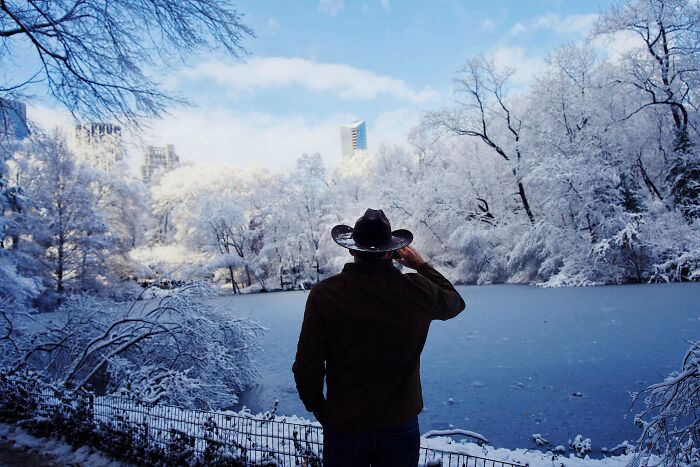 Man in a cowboy hat enjoying a cozy winter scene by a frozen lake surrounded by snow-covered trees and city buildings.