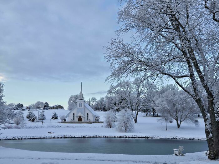 Snow-covered church and trees near a frozen pond, showcasing cozy winter scenes to enjoy without stepping into the cold.
