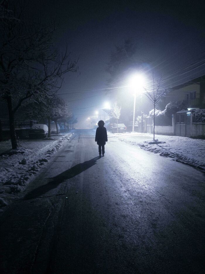 Silhouette of a person walking on a snowy street at night with streetlights glowing in the cozy winter scene.