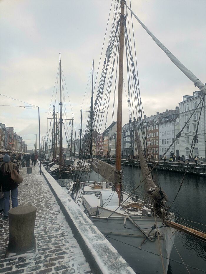 Cozy winter scene along a snowy canal with boats docked and people walking by colorful riverside buildings.