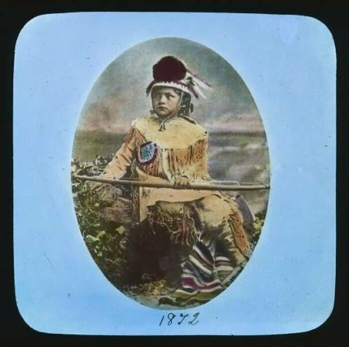 Vintage photo of a Native American child in traditional attire holding a spear, reflecting unsettling history and crime.