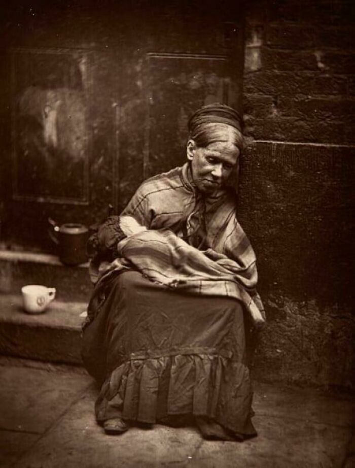 Victorian era woman sitting on a street corner, wrapped in a shawl, holding a bundled child in a sepia-toned photo.