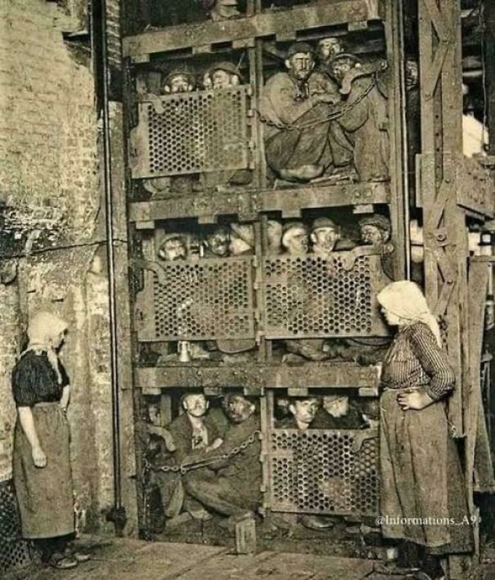 Victorian era coal miners crowded in a cage lift while women stand nearby in an industrial setting.