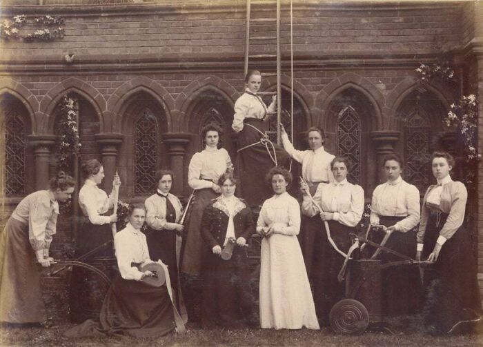 Group of Victorian era women in period clothing posing outdoors near brick building with arched windows and climbing ropes.