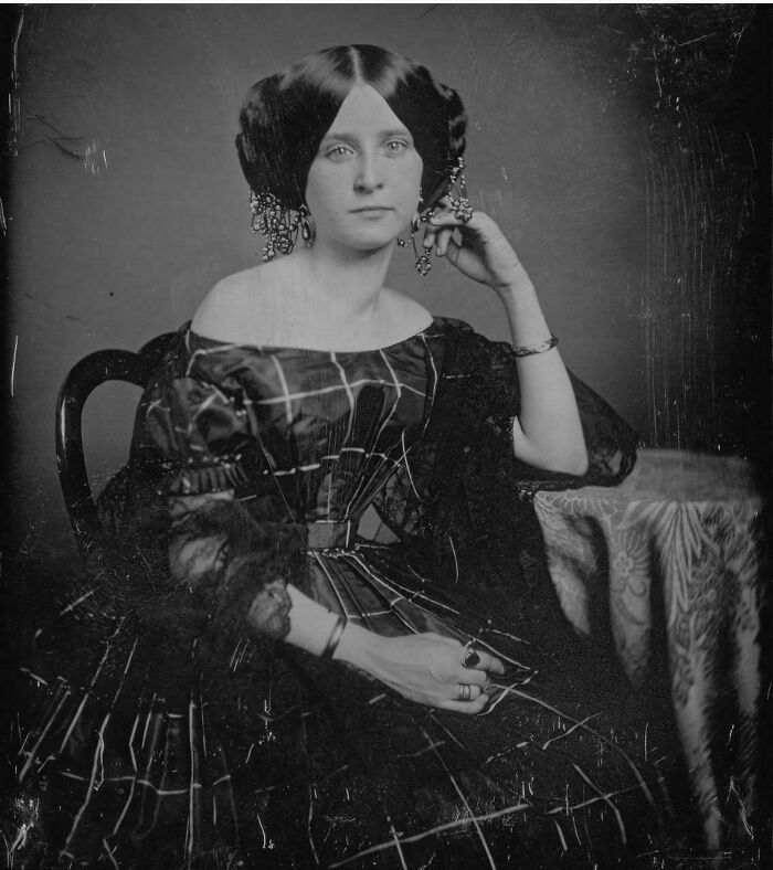 Victorian era woman in a plaid dress with elaborate hairstyle and jewelry sitting beside a patterned tablecloth.