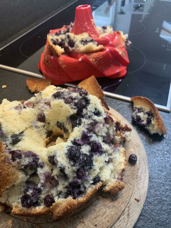 Blueberry cake ruined after baking failure, broken and misshapen on a wooden cutting board with a bent red bundt pan nearby.