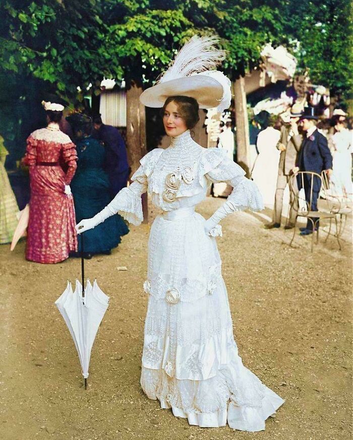 Victorian era woman in elegant white dress and large hat holding umbrella outdoors among other period-dressed people.