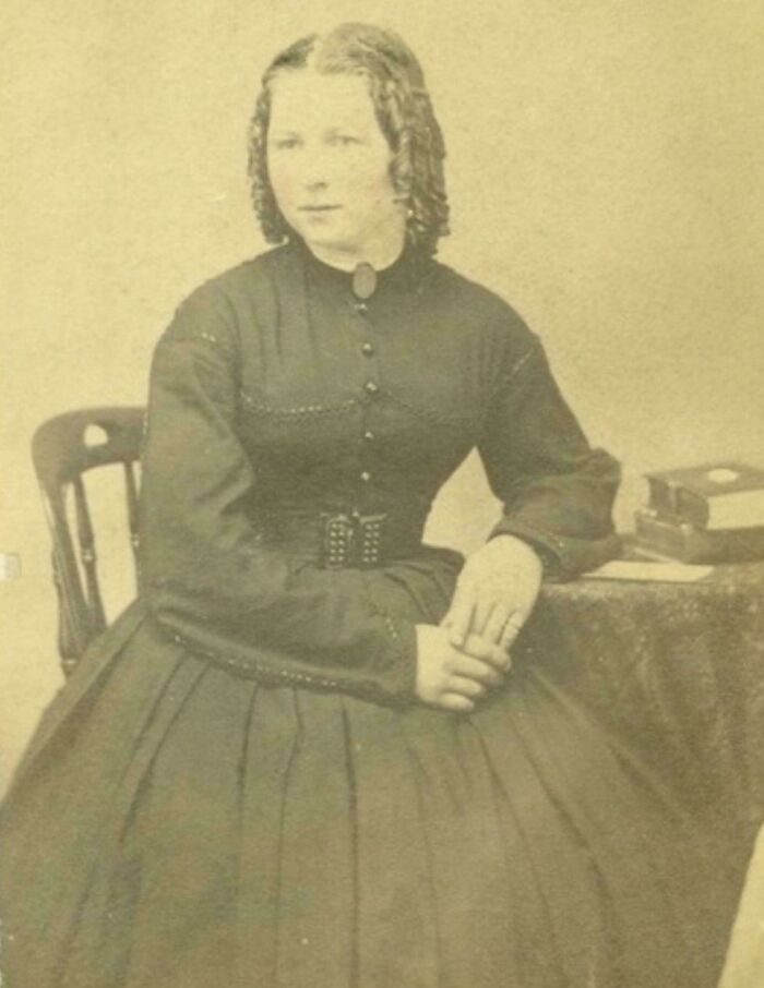 Victorian era woman in dark dress sitting beside a table with books, showcasing fashion from the different time period.