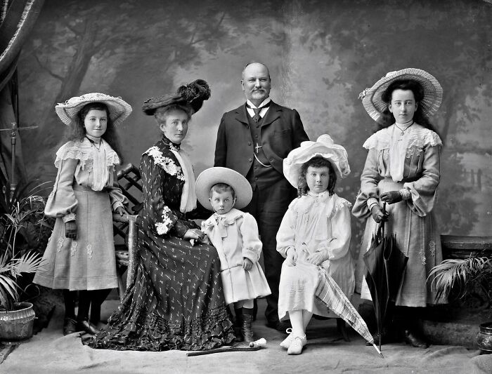 Victorian era family portrait with adults and children dressed in period clothing and large hats, holding umbrellas.